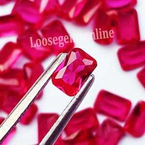 May include: A collection of vibrant red, rectangular-cut gemstones with faceted surfaces. One gemstone is held by metal tweezers in the foreground, while others are scattered behind it. The text Loosegemsonline is visible in white.
