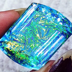 May include: A rectangular faceted gemstone featuring a vibrant play of color with deep blue, bright green, and golden yellow flashes. The stone has a polished surface with sharp edges and a clear, crystalline structure throughout the piece.