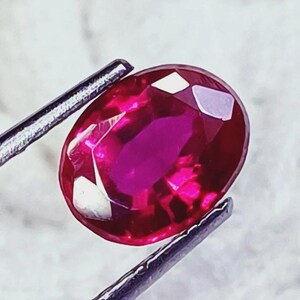 May include: A vibrant, oval-cut ruby gemstone with a deep red hue and multifaceted surface, held securely by a pair of metallic tweezers. The stone exhibits internal clarity and light reflection across its polished, symmetrical, and faceted edges.