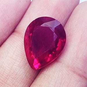 May include: A pear-shaped, faceted gemstone with a deep, vibrant red colour. The stone features a polished surface with multiple geometric facets that reflect light, creating a brilliant appearance across its teardrop-like silhouette.