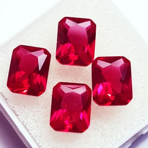 Puede incluir: Cuatro piedras preciosas rectangulares, facetadas y de color rojo intenso dispuestas en grupo. Cada piedra presenta una superficie clara y pulida con bordes afilados y un tono carmesí brillante y saturado que refleja la luz.