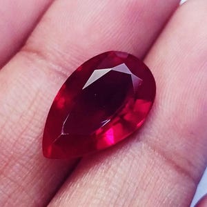Pode incluir: Uma pedra preciosa lapidada em forma de pera com uma cor vermelha profunda e vibrante. A pedra apresenta uma superfície polida com múltiplas facetas geométricas que refletem a luz, criando uma aparência brilhante. A pedra é translúcida e exibe um rico tom carmesim.