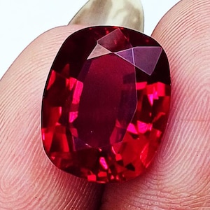 Puede incluir: Una piedra preciosa de talla cojín con un color rojo intenso y vibrante y una superficie facetada que refleja la luz. La piedra presenta una forma rectangular redondeada y simétrica con facetas internas geométricas y nítidas.
