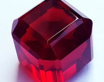 May include: A deep red, translucent, square-shaped gemstone with faceted edges. The object displays a high-gloss finish and sharp, geometric corners, reflecting light through its transparent body to create varying shades of crimson and ruby red.