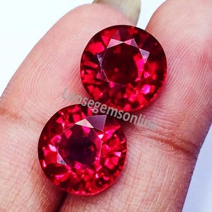 May include: Two round, faceted, vibrant red gemstones with a brilliant cut. The stones exhibit a deep crimson hue and reflective surfaces. The text "Loosegemsonline" is superimposed across the centre of the two circular, polished red gems.