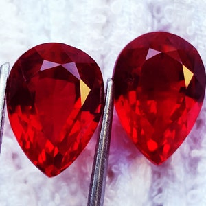 May include: Two vibrant, pear-shaped red gemstones with faceted surfaces that reflect light. The stones are held by silver-toned metal tweezers, showcasing their deep crimson colour and precise cut. The gems are positioned side by side, displaying clarity.