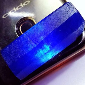 May include: A vibrant, rectangular, deep blue crystal with a faceted surface. The crystal is displayed on a reflective surface, showcasing its rich color and natural texture. The crystal's shape suggests it is a raw, uncut gemstone.