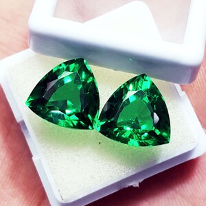 May include: Two vibrant green, trillion-cut gemstones with faceted surfaces displayed inside a small, white, square plastic storage box with a clear lid. The stones exhibit a deep, saturated emerald hue and are positioned on a white foam insert.