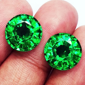 Puede incluir: Dos gemas redondas, facetadas, de color verde brillante. Las gemas se sostienen entre los dedos sobre un fondo blanco. Las gemas tienen un corte brillante, que refleja la luz y crea un efecto brillante. Las gemas son de un verde vibrante y llamativo.