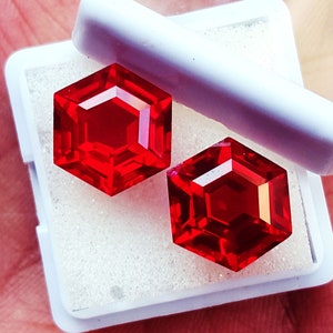 Könnte beinhalten: Zwei leuchtend rote, sechseckig geschliffene Edelsteine mit facettierten Oberflächen befinden sich in einer kleinen, weißen, quadratischen Aufbewahrungsbox. Die Steine liegen auf einem weißen Schaumstoffeinsatz und zeigen ihre tiefrote Farbe.