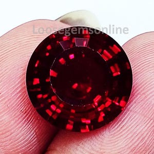 Puede incluir: Una piedra preciosa redonda y facetada de color rojo intenso con un corte brillante, que muestra reflejos de luz internos y un tono carmesí oscuro. La piedra presenta una tabla circular y múltiples facetas triangulares en el perímetro, con el texto Loosegemsonline visible.