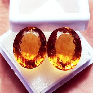 Könnte beinhalten: Zwei oval geschliffene Citrin-Edelsteine mit einem leuchtend gold-orangen Farbton und facettierten Oberflächen. Die Steine liegen nebeneinander in einer weißen Schmuckschatulle aus Kunststoff mit einer weich gepolsterten Innenauskleidung.