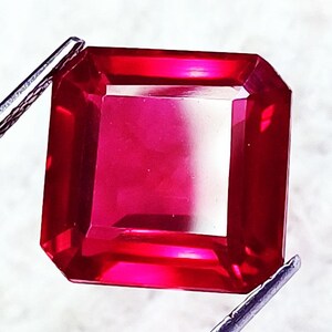 May include: A square-cut, vibrant red gemstone with faceted surfaces that reflect light, held by metal tweezers. The stone displays a deep, saturated crimson hue with clear transparency and sharp, defined edges throughout its geometric shape.