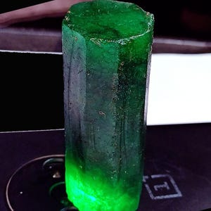 May include: A raw, hexagonal emerald crystal with a deep green color and a translucent, glowing base. The gemstone features a vertical, columnar structure with natural surface textures, facets, and internal inclusions visible throughout the stone.