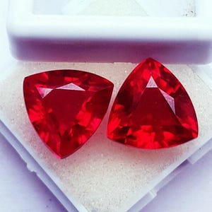 May include: Two vibrant red gemstones cut into a triangular shape with multiple facets. The stones are displayed side by side, showcasing a deep crimson hue and a brilliant reflective surface, resting within a white container with a textured base.
