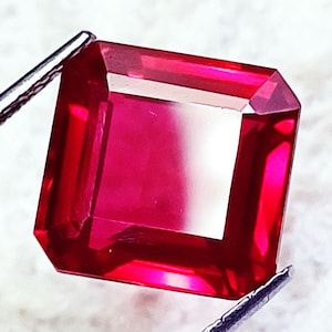 May include: A square-cut, vibrant red gemstone with faceted surfaces that reflect light. The transparent stone is held by metallic tweezers at the top left and bottom right corners, showcasing its deep crimson colour and clear, polished clarity.