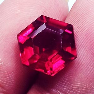 Könnte beinhalten: Ein leuchtend tiefroter Edelstein in präziser sechseckiger Form. Der Stein weist mehrere facettierte Oberflächen auf, die das Licht reflektieren und ein brillantes, durchscheinendes Aussehen mit scharfen, definierten Kanten erzeugen.