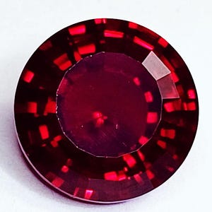 Könnte beinhalten: Ein runder, facettierter Edelstein in einer tiefen, leuchtend roten Farbe. Der Stein weist einen komplexen, geometrischen Schliff mit einer zentralen runden Tafel auf, die von mehreren konzentrischen Reihen rechteckiger Facetten umgeben ist.