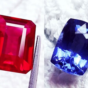 May include: Two gemstones, a red ruby and a blue sapphire, are displayed on a white surface. The ruby is a rectangular emerald cut, while the sapphire is a cushion cut.