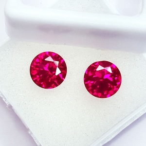 May include: Two round, bright pink gemstones in a white box. The gemstones are faceted and have a high shine.