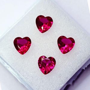 Puede incluir: Cuatro piedras preciosas facetadas en forma de corazón de un vibrante color rojo magenta. Cada piedra presenta un corte brillante con múltiples superficies reflectantes, dispuestas en forma de diamante sobre una almohadilla de espuma blanca texturizada.