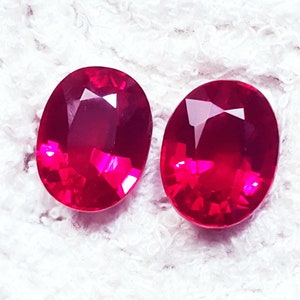 Puede incluir: Dos piedras preciosas de color rojo intenso con corte ovalado y superficies facetadas que reflejan la luz. Estas piedras vibrantes y translúcidas presentan una forma simétrica y un acabado pulido, mostrando un rico color carmesí.