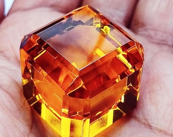 May include: A faceted, transparent, amber-colored gemstone cut into a cube shape. The stone features sharp, geometric edges and polished surfaces that reflect light, creating deep orange and yellow highlights throughout the crystalline structure.