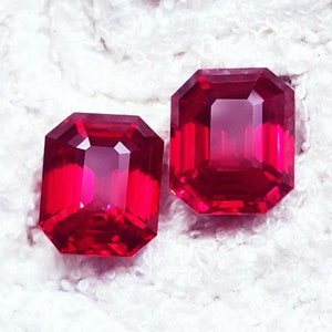 May include: Two radiant, rectangular-cut, deep red gemstones. The gems have multiple facets, reflecting light and creating a brilliant sparkle. The stones are set against a white, textured background, highlighting their vibrant color and clarity.
