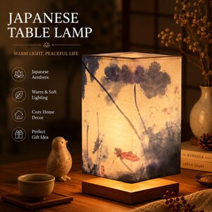 May include: A rectangular table lamp with a wooden base and a fabric shade featuring a blue ink-wash style painting of lotus leaves and a single orange fish. The lamp emits a warm glow and includes text reading JAPANESE TABLE LAMP.