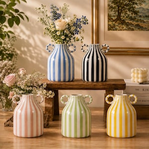 Pode incluir: Cinco vasos de cerâmica com duas alças laterais circulares e listras verticais em azul, preto, rosa, verde e amarelo. O vaso azul contém um buquê de rosas brancas e flores azuis. Cada vaso apresenta um corpo arredondado e pescoço.