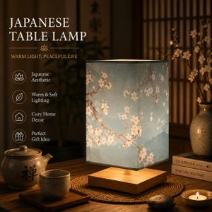 May include: A square table lamp with a light wood base and a fabric shade featuring a blue background with white cherry blossom branches. The lamp emits a warm glow and includes text on the left side reading JAPANESE TABLE LAMP.