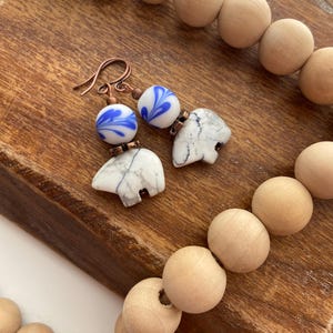 May include: A pair of earrings with white and blue ceramic beads and copper wire. The beads have a marbled pattern and a blue floral design. The earrings are hanging from a wooden surface.