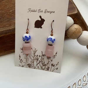 May include: A pair of dangle earrings with a pink and white layered bead and a blue and white porcelain bead. The earrings are on a white card with the text "Rabbit Ear Designs" and a silhouette of a rabbit.