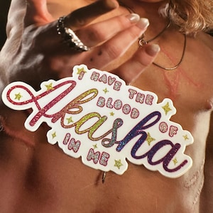 May include: A white die-cut sticker featuring the glittery, multi-colored text "I HAVE THE BLOOD OF Akasha IN ME" with small yellow star accents. The sticker is placed over a magazine cover showing a person with long hair and a silver necklace.