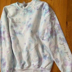 May include: A tie-dye sweatshirt in light blue, purple, and white. A silver glitter snowflake design is on the front. The right sleeve has the words "Step into your power" in silver glitter. The sweatshirt has a crew neck and long sleeves.