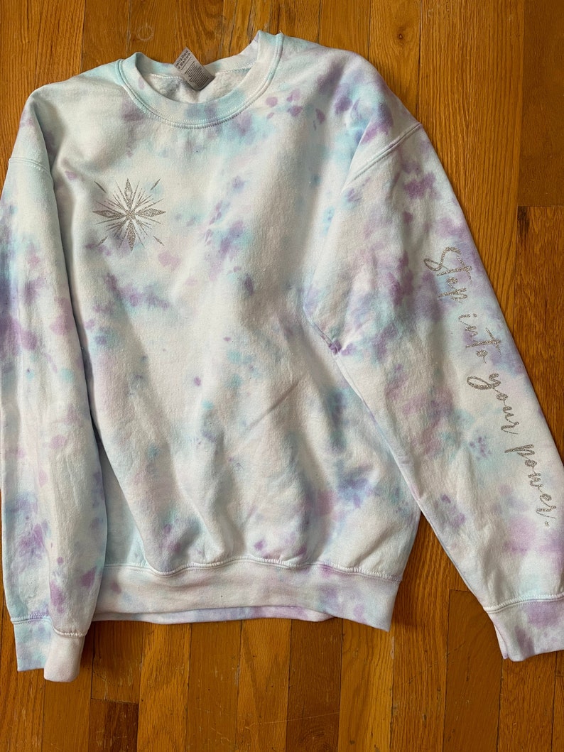 May include: A tie-dye sweatshirt in light blue, purple, and white. A silver glitter snowflake design is on the front. The right sleeve has the words "Step into your power" in silver glitter. The sweatshirt has a crew neck and long sleeves.