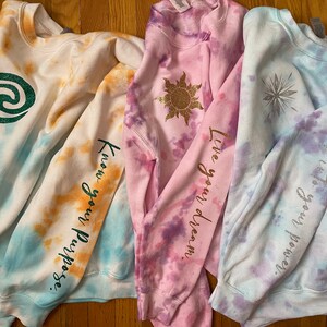 May include: Four tie-dye sweatshirts in white, blue, orange, and pink. Each sweatshirt has a graphic and text on the sleeve. The graphics include a green swirl, a gold sun, and a silver snowflake. The text includes "Know your Purpose", "Live your dream", and "She is your power."