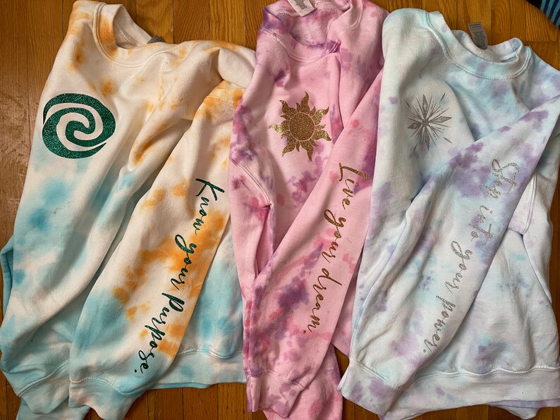 May include: Four tie-dye sweatshirts in white, blue, orange, and pink. Each sweatshirt has a graphic and text on the sleeve. The graphics include a green swirl, a gold sun, and a silver snowflake. The text includes "Know your Purpose", "Live your dream", and "She is your power."