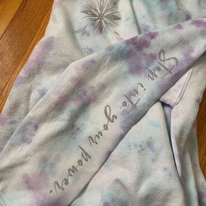 May include: A tie-dye sweatshirt in light blue, purple, and white. The left sleeve has the text "Step into your power" in silver glitter. A silver glitter snowflake design is on the upper left chest.
