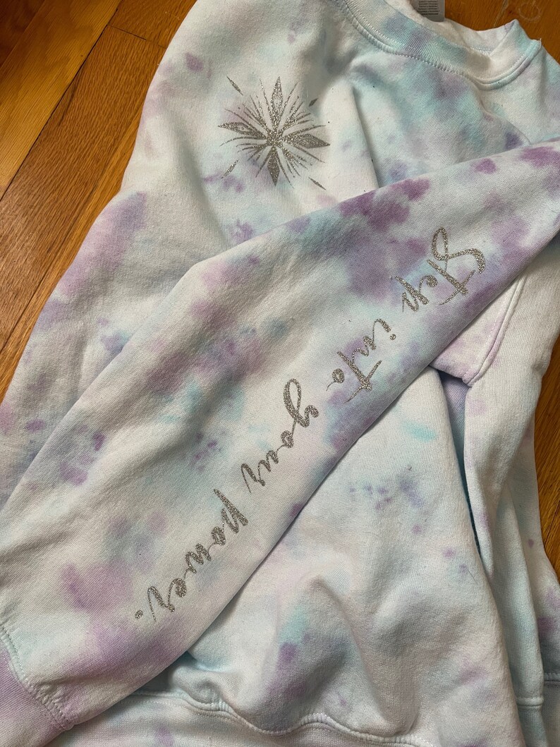 May include: A tie-dye sweatshirt in light blue, purple, and white. The left sleeve has the text "Step into your power" in silver glitter. A silver glitter snowflake design is on the upper left chest.