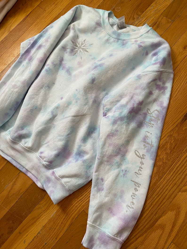 May include: A crew neck sweatshirt with a tie-dye pattern in pale blue, lilac, and white. It features a silver glitter snowflake design and the text "Step into your power" on the right sleeve. The cuffs and hem are ribbed.
