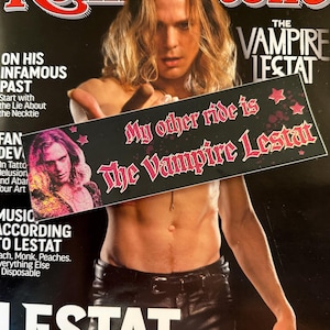 May include: A rectangular bumper sticker with a black background featuring the pink text 'My other ride is The Vampire Lestat' in a gothic font, accented with pink stars and a small portrait of a long-haired individual on the left side.