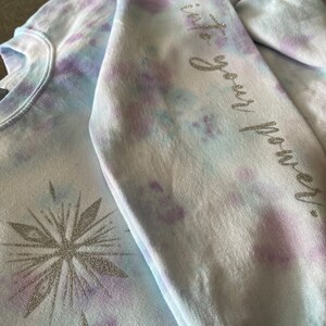 May include: A tie-dye sweatshirt in blue, purple, and white. The left side has a silver glitter snowflake design. The right sleeve features the phrase "Step into your power" in silver glitter lettering. The sweatshirt has a crew neck.