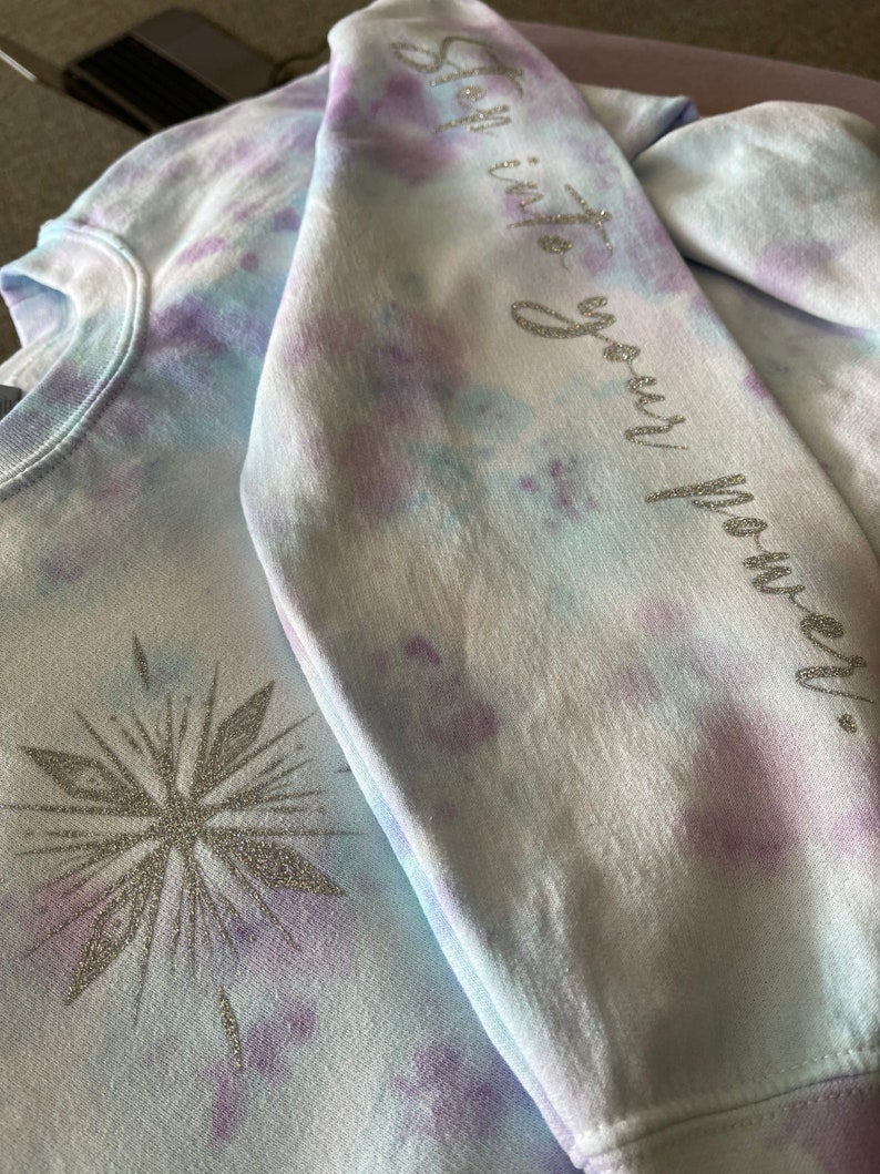 May include: A tie-dye sweatshirt in blue, purple, and white. The left side has a silver glitter snowflake design. The right sleeve features the phrase "Step into your power" in silver glitter lettering. The sweatshirt has a crew neck.