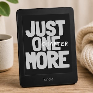 May include: A black e-reader device with a rectangular screen displaying the white text JUST ONE MORE CHAPTER in a bold, sans-serif font. The device features a thin black bezel and the word kindle printed in small white letters at the bottom.