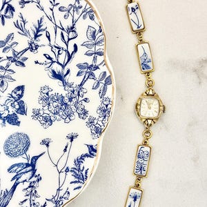 May include: A gold-tone wristwatch with a square face and a link bracelet featuring rectangular white ceramic tiles decorated with blue floral and hummingbird motifs, positioned next to a white plate with a matching blue and white botanical pattern.