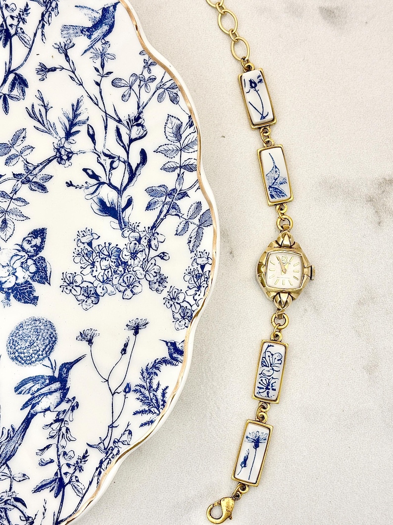 May include: A gold-tone wristwatch with a square face and a link bracelet featuring rectangular white ceramic tiles decorated with blue floral and hummingbird motifs, positioned next to a white plate with a matching blue and white botanical pattern.