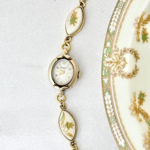 Puede incluir: Un reloj vintage dorado con esfera blanca y la marca "GANT". La correa del reloj presenta eslabones ovalados blancos con dise&ntilde;os florales y detalles dorados. Tambi&eacute;n se ve un plato decorativo con motivos florales.