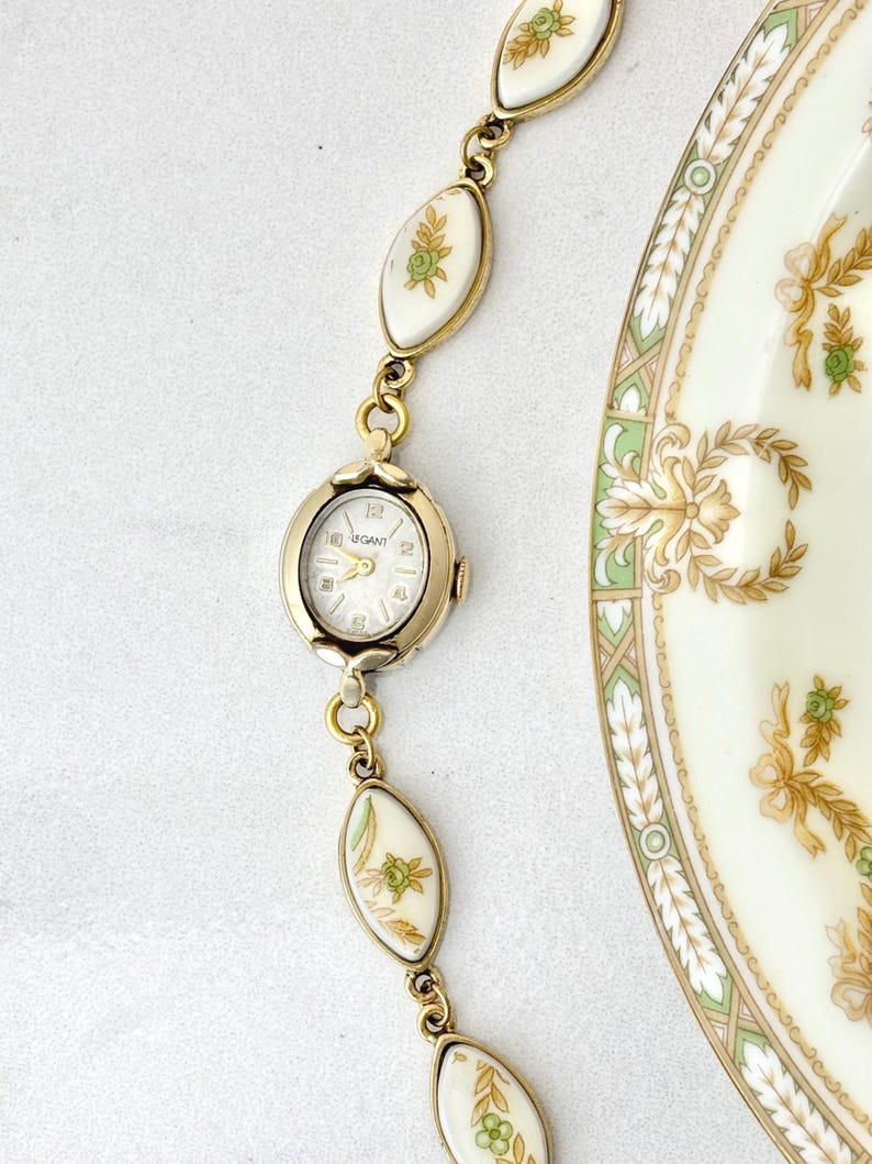 Puede incluir: Un reloj vintage dorado con esfera blanca y la marca "GANT". La correa del reloj presenta eslabones ovalados blancos con dise&ntilde;os florales y detalles dorados. Tambi&eacute;n se ve un plato decorativo con motivos florales.