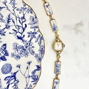 May include: A gold-tone link bracelet watch featuring rectangular ceramic tiles with blue and white floral and hummingbird patterns. The watch has a square face with gold hands and markers, connected by gold metal links and a lobster clasp closure.